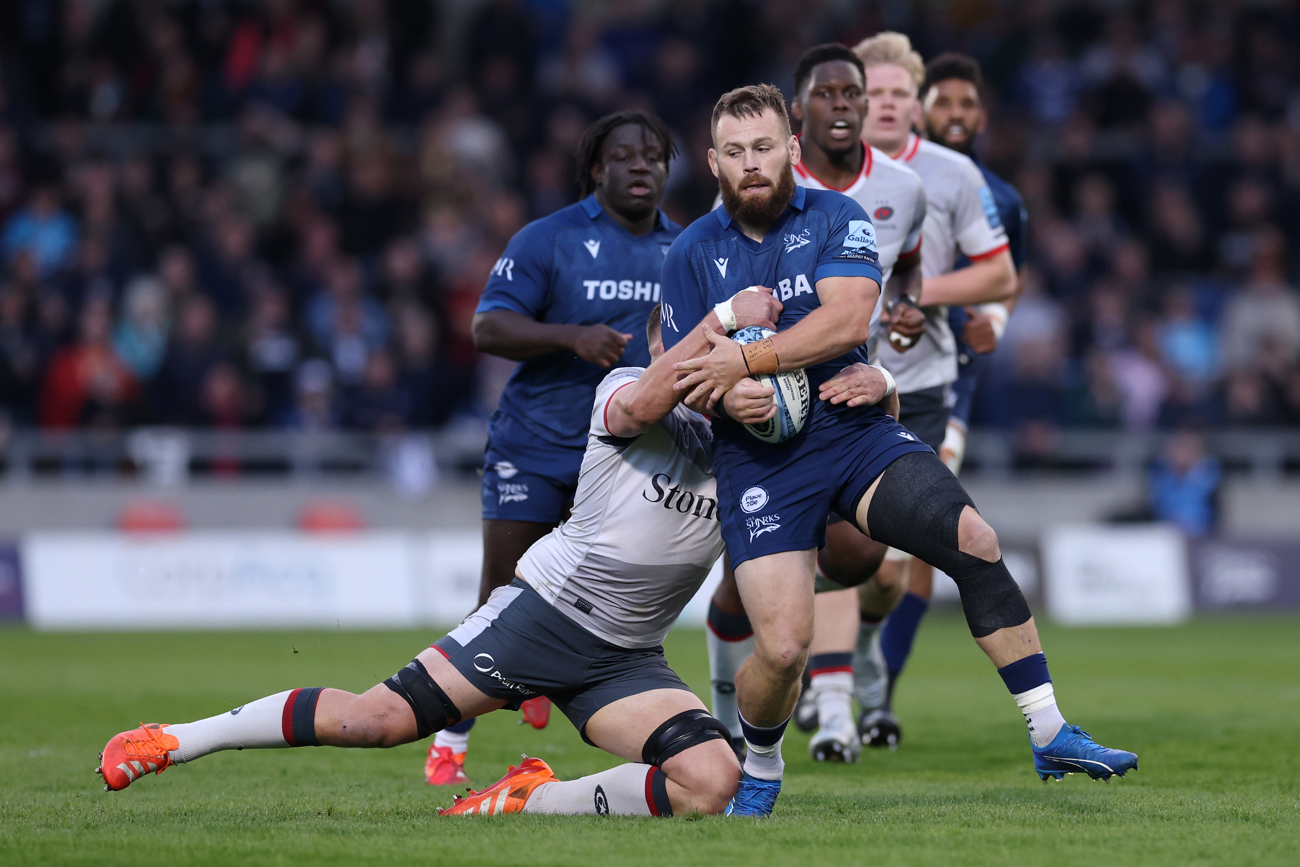A rugby player from Sale Sharks is tackled by a Saracens player.